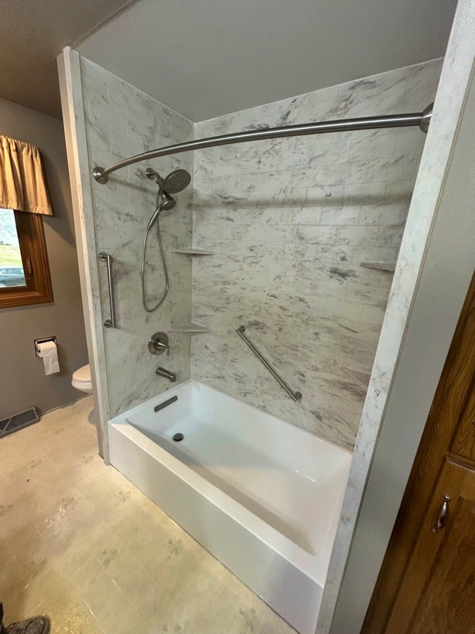 Bathtub replacement with a white bathtub, grey and white marble look walls, a brushed nickel rain shower head and handheld, a grab bar, corner shelves and a curved curtain rod in a Wisconsin home