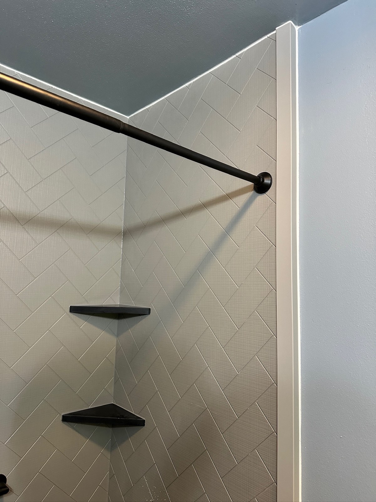 Detail of grey herringbone textured shower walls with black corner shelves and a matte black curtain rod in Weyauwega, WI