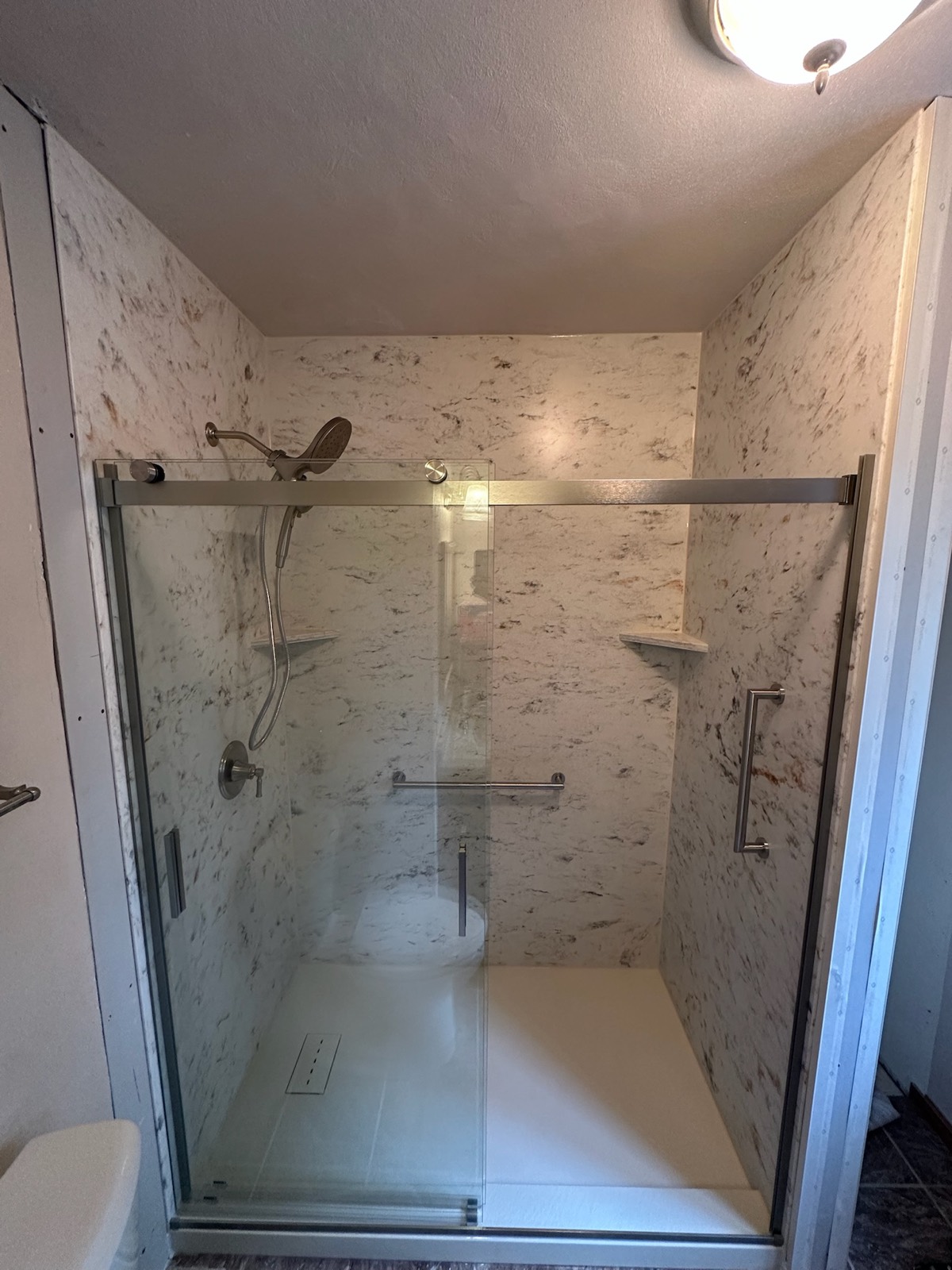 Shower with cream and brown veined marble look walls, a sliding glass door, a brushed nickel shower head with handheld, grab bars, corner shelves and a low threshold base in Neenah, WI