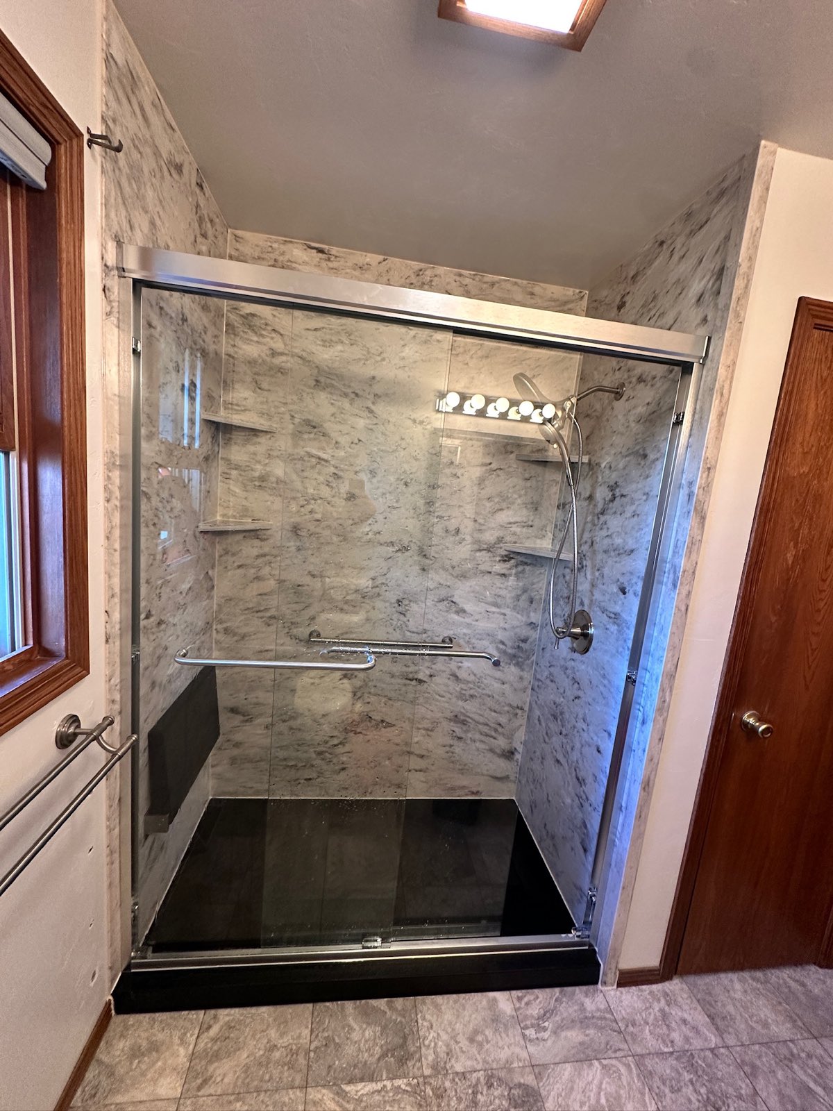 Walk in shower with grey marble look walls, a sliding glass door, a handheld shower on a slide bar, brushed nickel grab bars, corner shelves, a black fold down seat and a black shower base in Spruce, WI