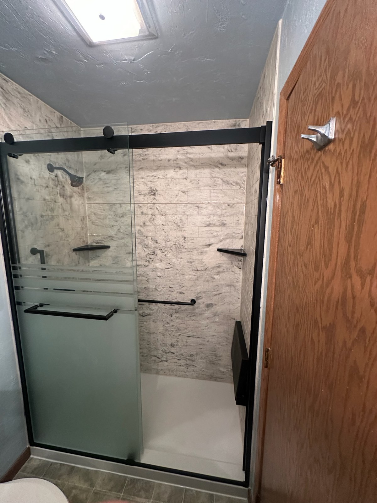 Shower with white and grey marble look walls, a black framed sliding glass door with a frosted band, a matte black shower head, black corner shelves, a black grab bar and a black fold down seat in Wausau, WI