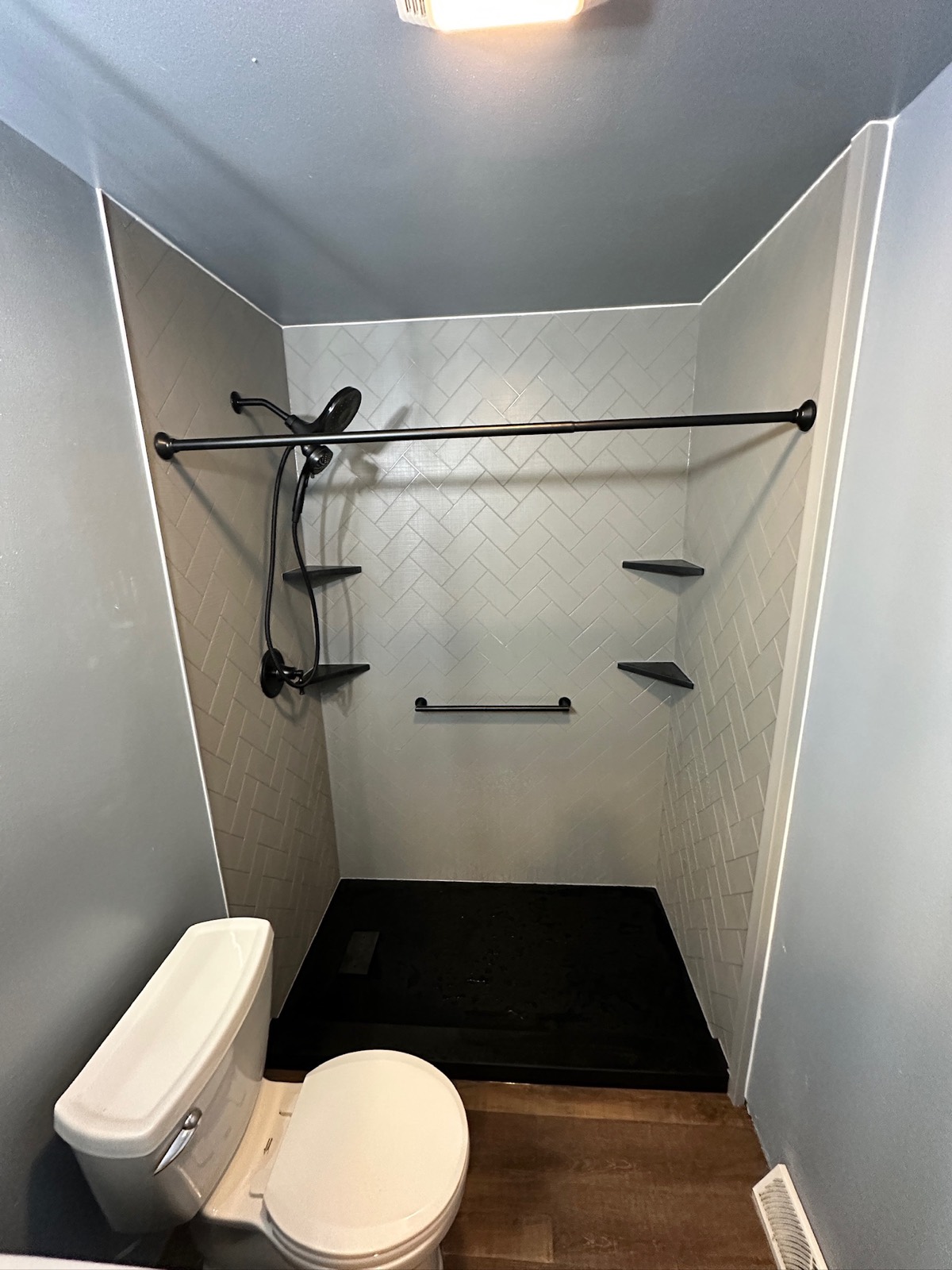 Low threshold shower with grey herringbone textured walls, a matte black rain shower head and handheld, black corner shelves, a black grab bar and a black shower base in Weyauwega, WI