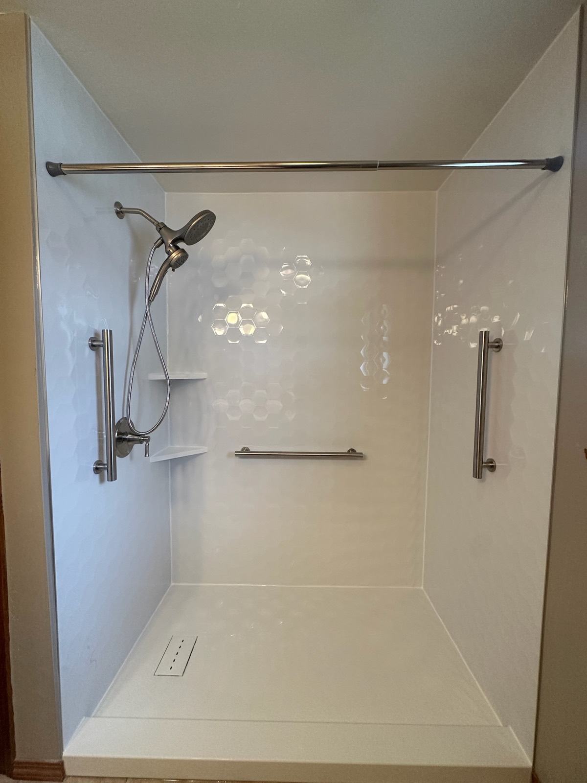 Close view of a tub to shower conversion with gloss white hexagon walls, grab bars on three walls, a handheld shower on a slide bar and corner shelves in Bellevue, WI