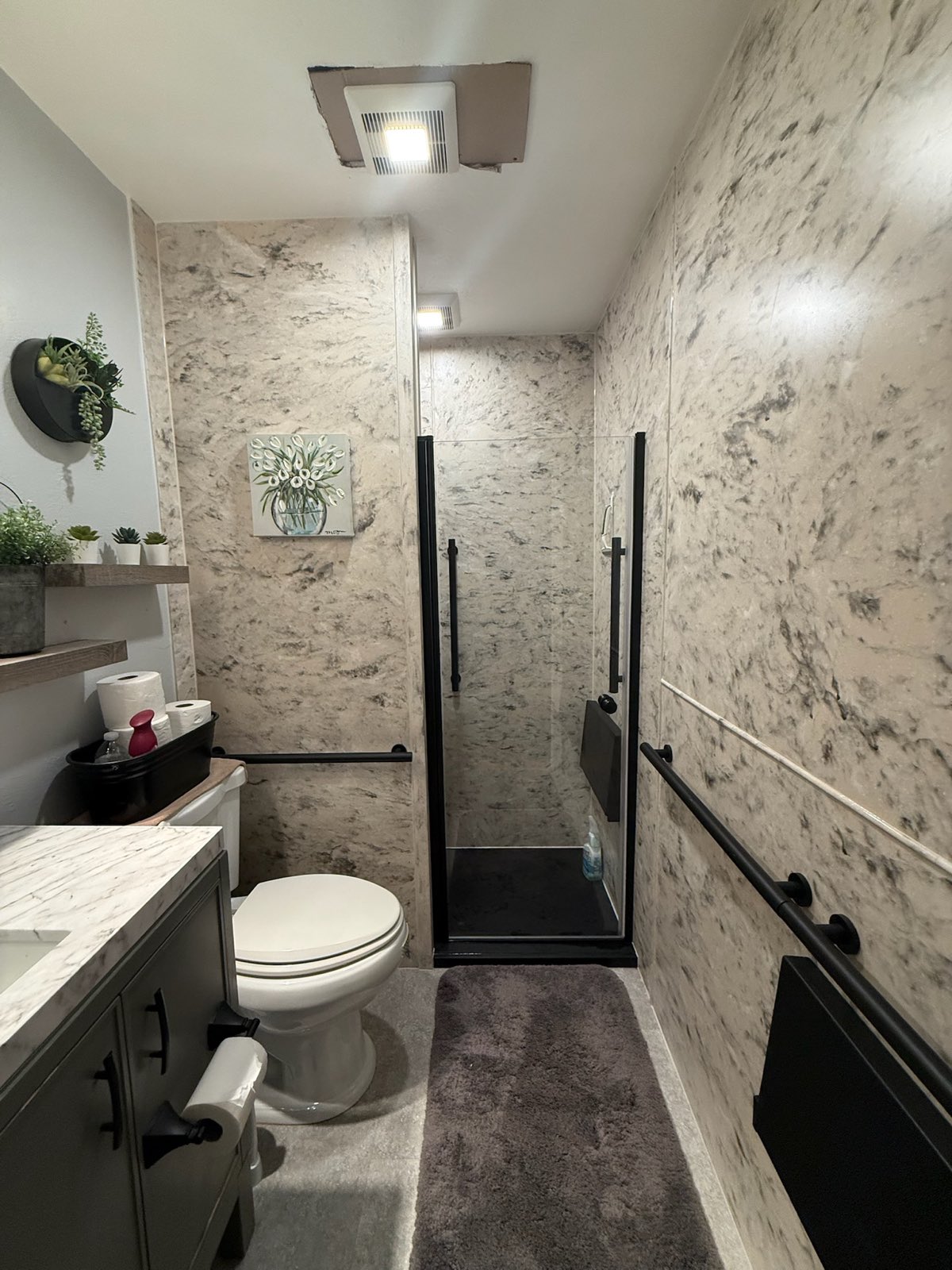 Grey veined marble look walls run through the whole bathroom around a black framed glass shower with black grab bars, a black fold down seat and a black shower base in Appleton, WI