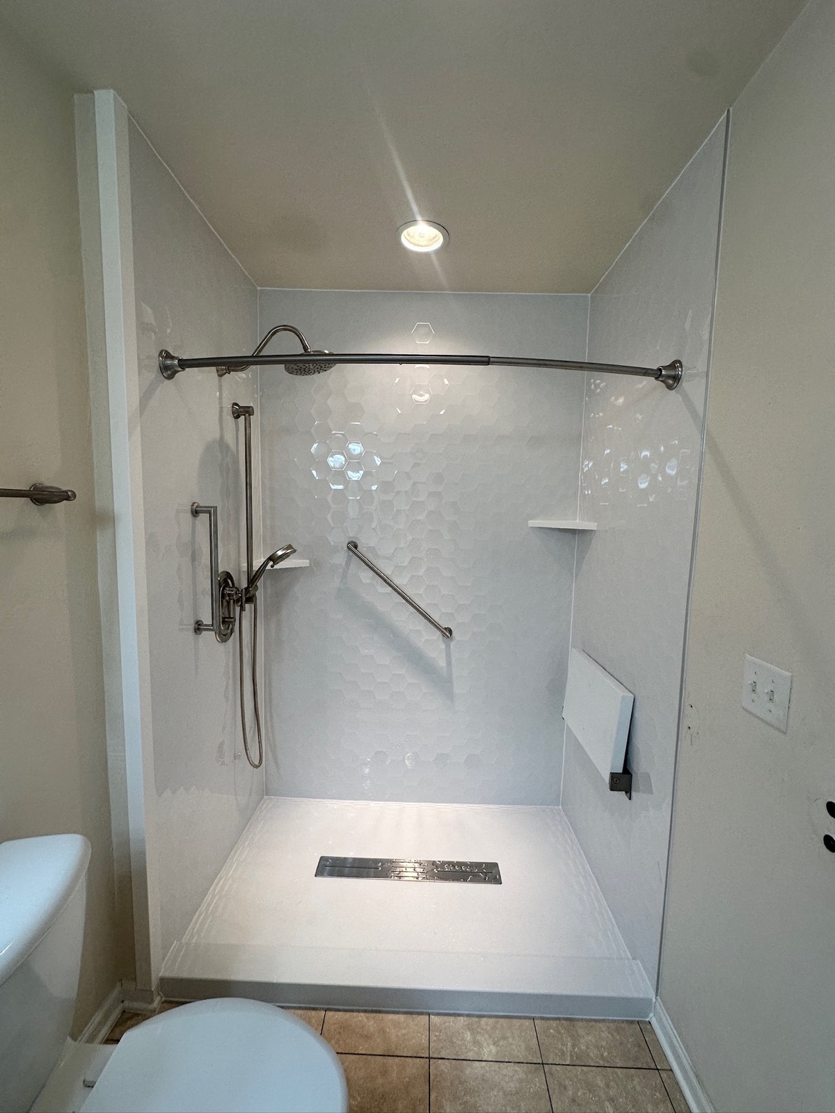 Walk in shower with gloss white hexagon walls, a rain shower head, a handheld shower on a slide bar, a fold down seat, grab bars, corner shelves and a linear drain in Appleton, WI