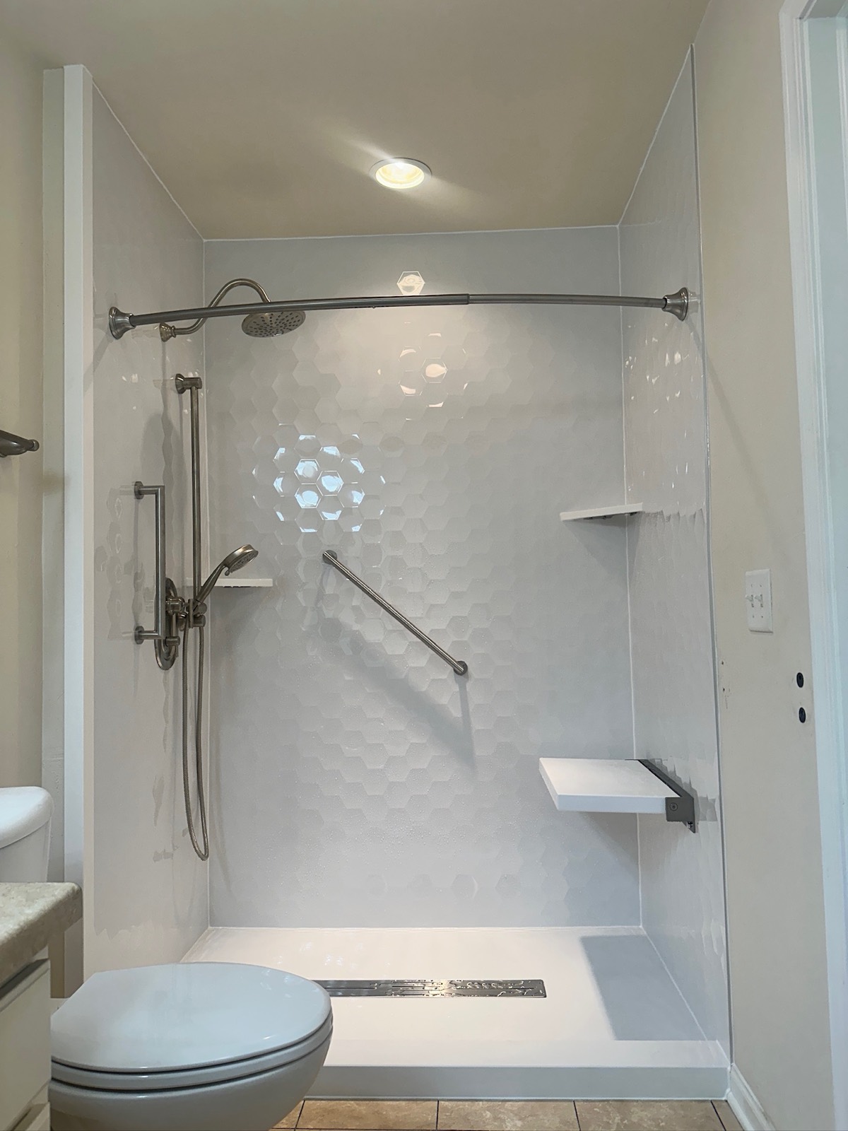 Walk in shower with Samuel Mueller Gloss White Hexagon walls, a brushed nickel rain shower head, a handheld shower on a slide bar, grab bars, a white fold down seat, corner shelves and a linear drain in Appleton, WI