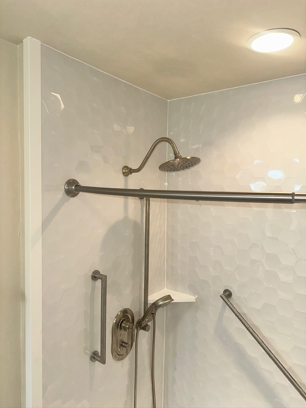 Detail of a brushed nickel rain shower head, handheld shower, grab bars and a corner shelf against gloss white hexagon shower walls in Appleton, WI