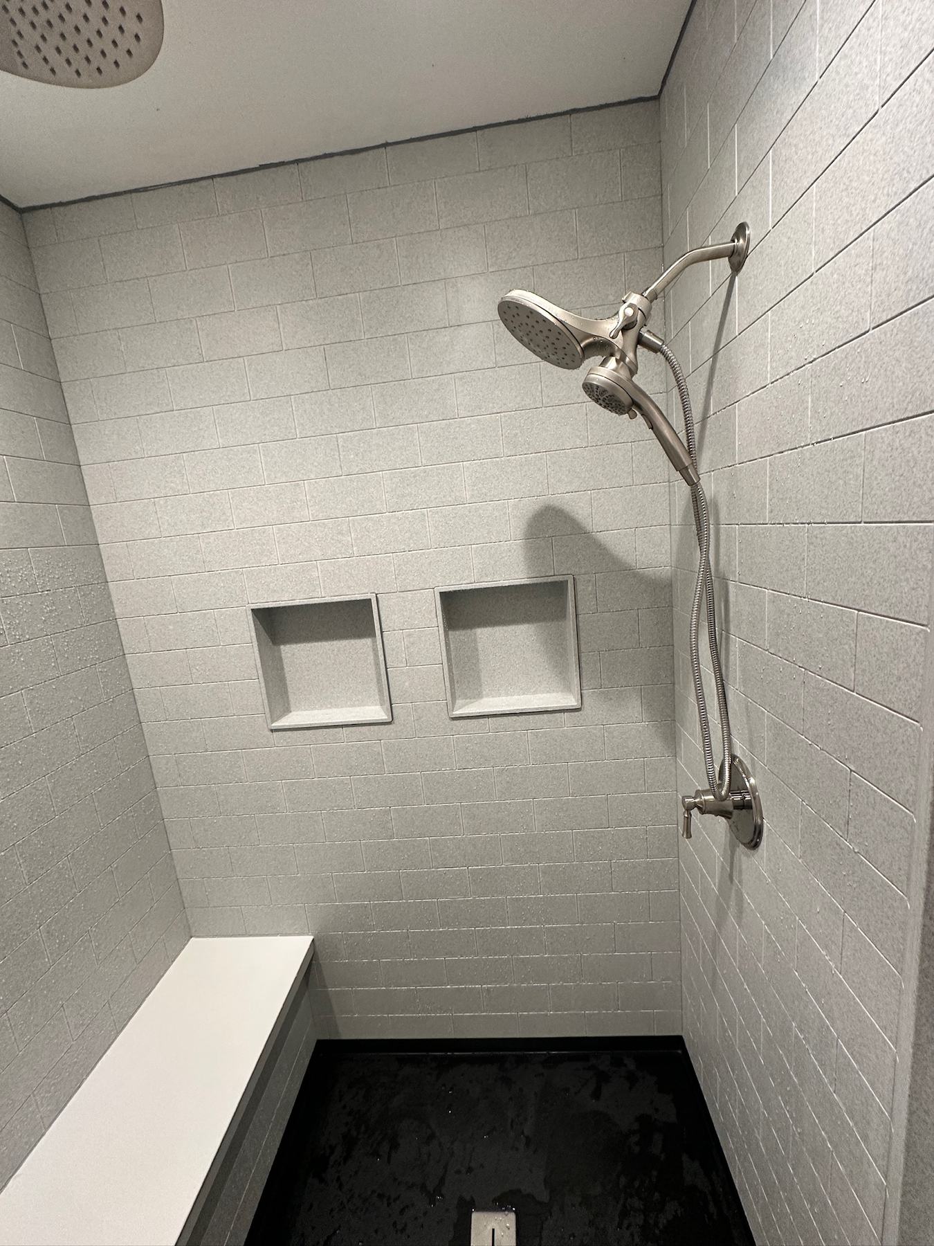 Walk in shower with grey textured subway look walls, a ceiling rain shower head, a brushed nickel shower head with handheld, two recessed niches, a bench seat and a black shower floor in Howard, WI