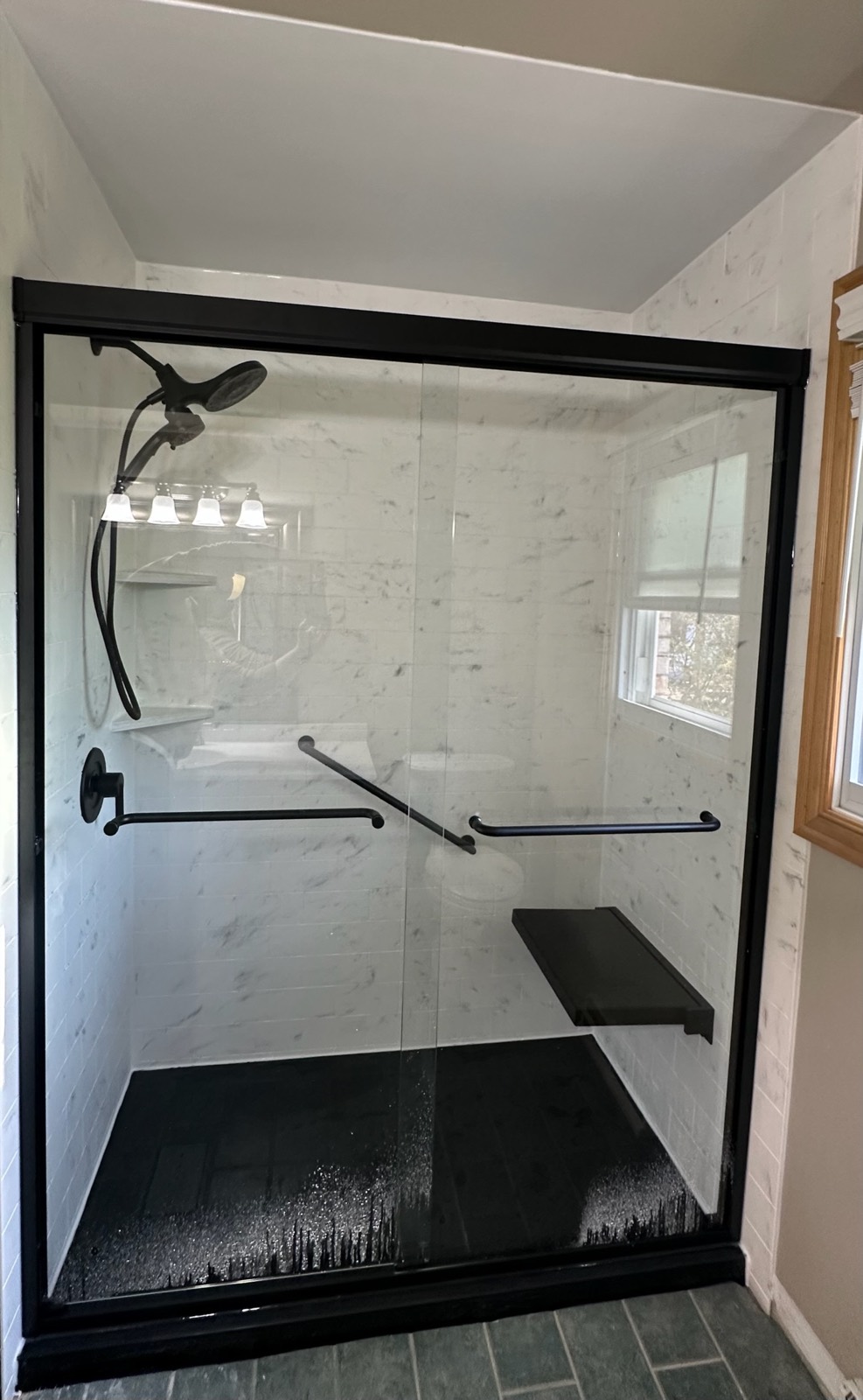 Walk in shower with white marble look walls, a black framed sliding glass door, black grab bars, a black fold down seat, a matte black shower head and a black shower base in Little Chute, WI