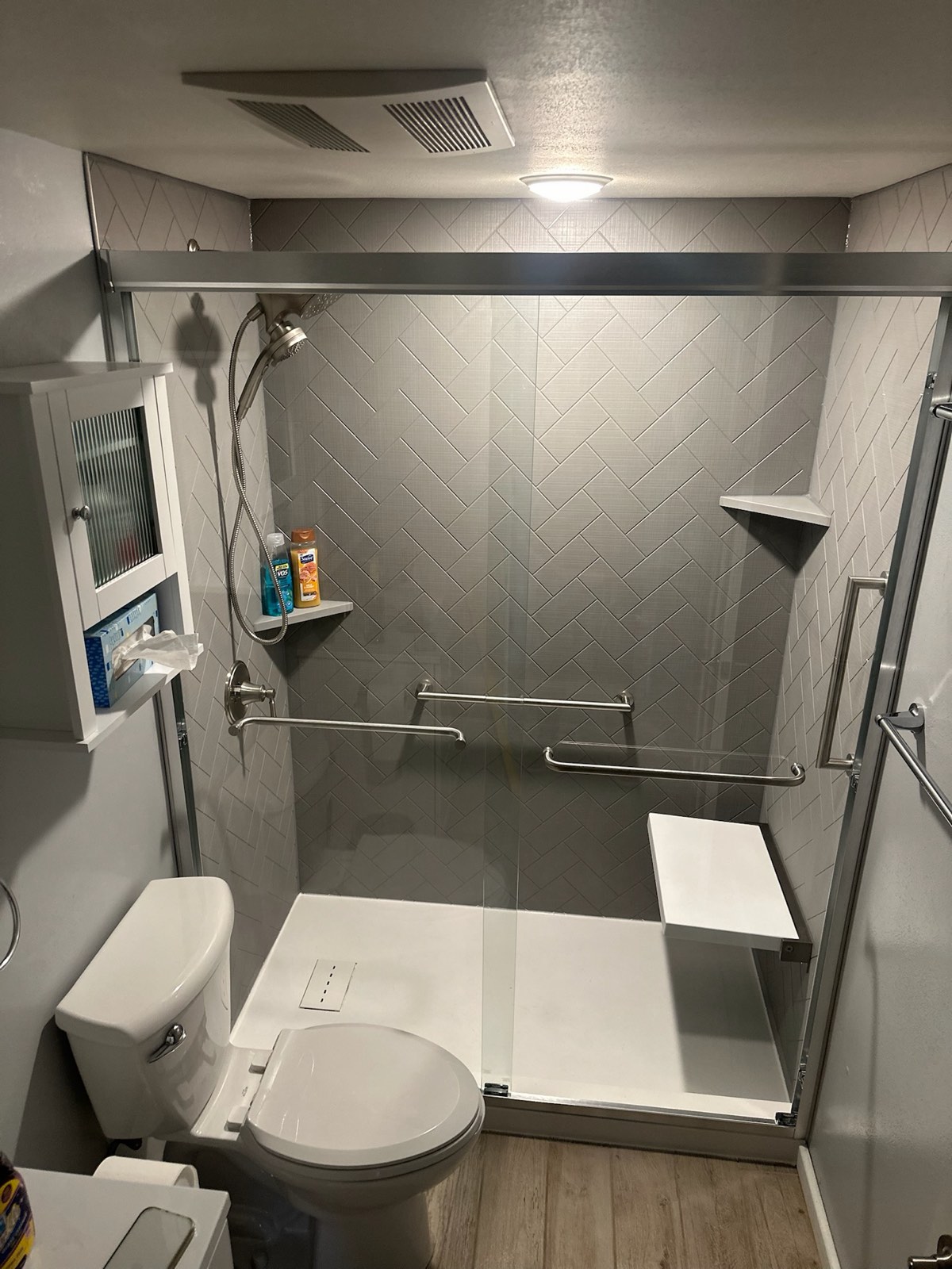Walk in shower with grey herringbone textured walls, a sliding glass door, brushed nickel grab bars, a white fold down seat, a handheld shower on a slide bar and a corner shelf in Marion, WI