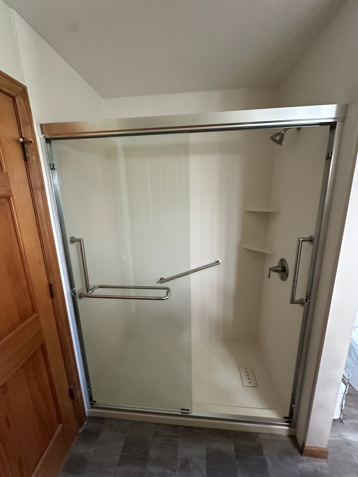 Walk in shower with smooth almond colored walls, a sliding glass door with one frosted panel, brushed nickel grab bars, corner shelves and a low threshold base in Pensaukee, WI