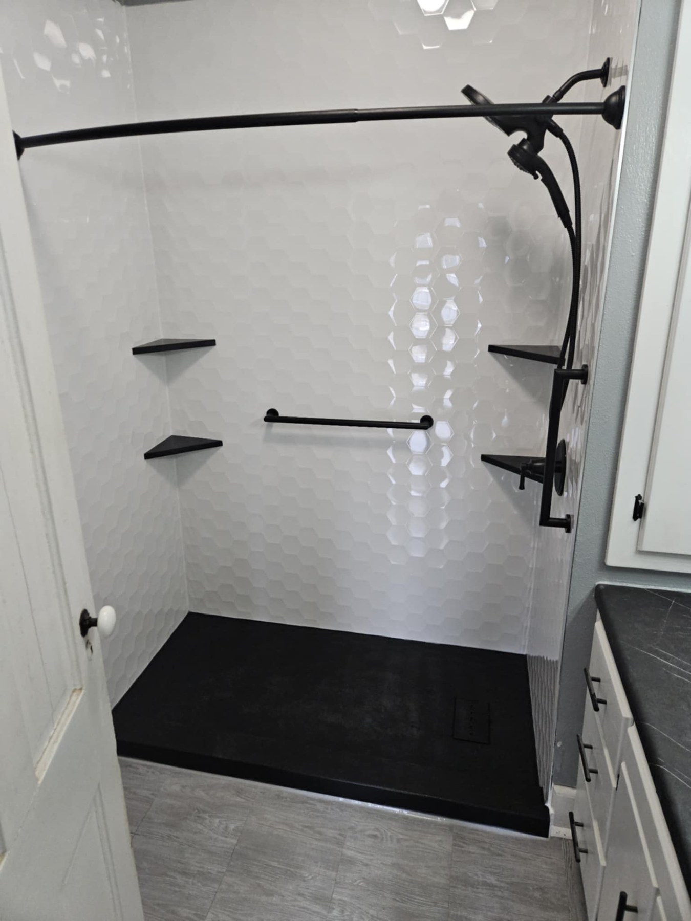 Shower with Samuel Mueller Gloss White Hexagon walls, a black shower base, black corner shelves, a black grab bar and a matte black handheld shower installed by American Shower Company in a Wisconsin home