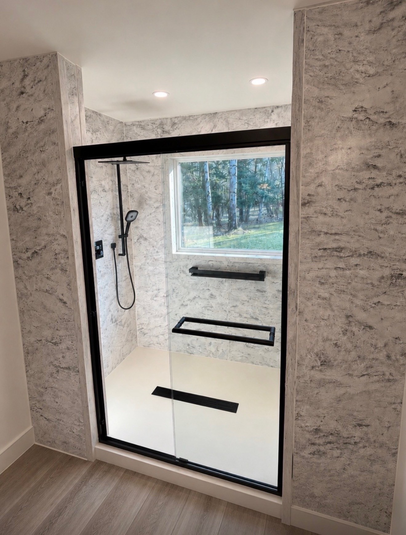 Shower replacement with grey marble look walls, a black framed sliding glass door, a window inside the shower, a matte black rain shower head and handheld, black grab bars, a white low threshold base and a linear drain installed by American Shower Company in a Wisconsin home