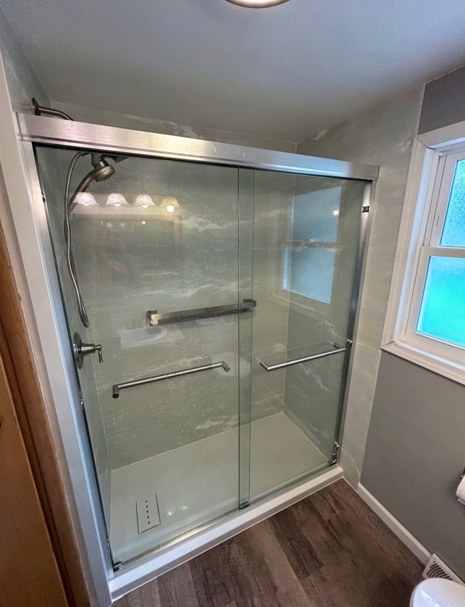 Shower with grey marble look walls, a chrome framed sliding glass door, grab bars, a handheld shower and a window beside the shower installed by American Shower Company in a Wisconsin home