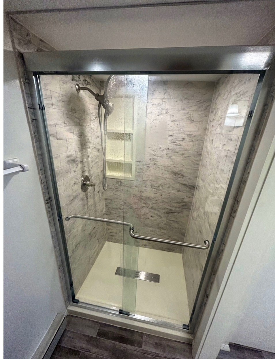 Shower with grey marble look walls, a chrome framed sliding glass door, a recessed niche, a grab bar, a handheld shower, a white base and a linear drain installed by American Shower Company in a Wisconsin home