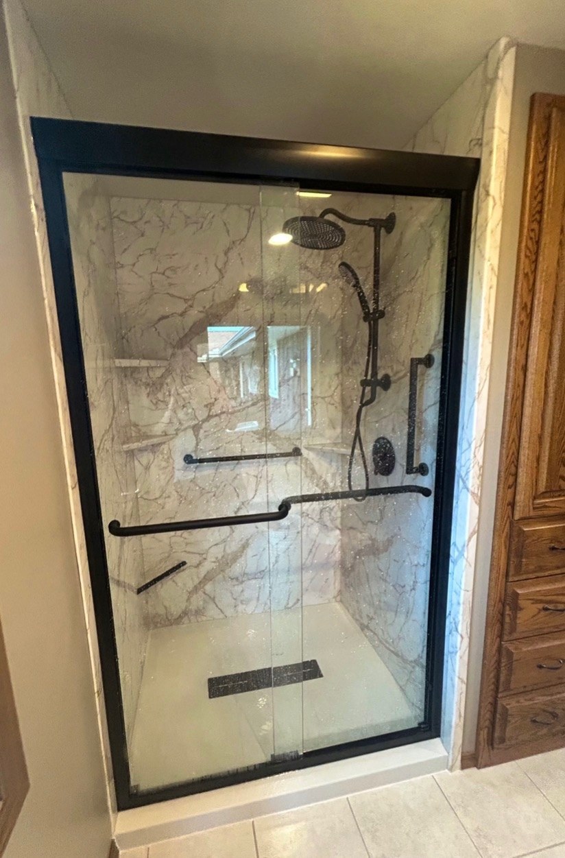 Shower with white walls, a black framed sliding glass door, black grab bars, a window inside the shower and a black shower base installed by American Shower Company in a Wisconsin home