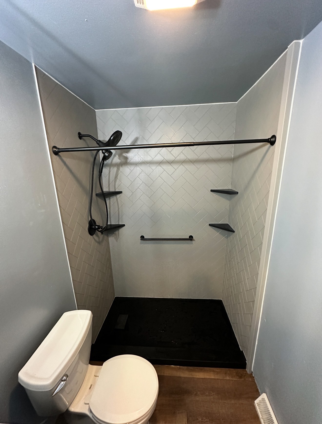 Shower with white herringbone textured walls, a black curtain rod, a matte black rain shower head and handheld, black corner shelves, a black grab bar and a black shower base installed by American Shower Company in a Wisconsin home