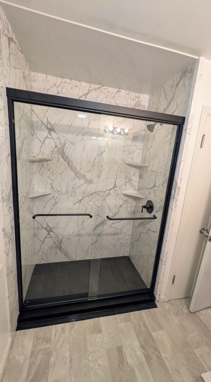 Shower replacement with white marble look walls, a black framed sliding glass door, black grab bars, corner shelves and a black shower base installed by American Shower Company in a Wisconsin home
