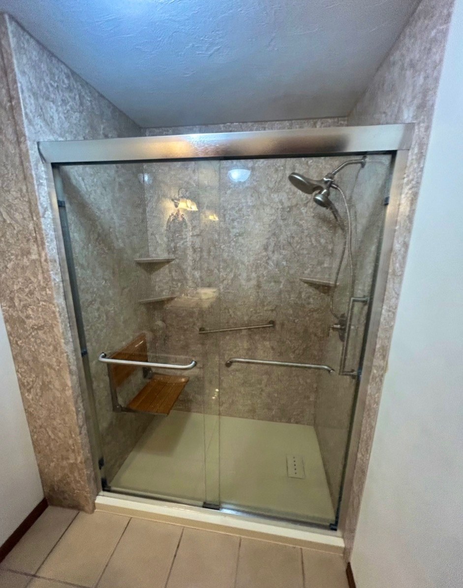 Shower with tan stone look walls, a chrome framed sliding glass door, a teak fold down seat, grab bars, corner shelves and a white base installed by American Shower Company in a Wisconsin home