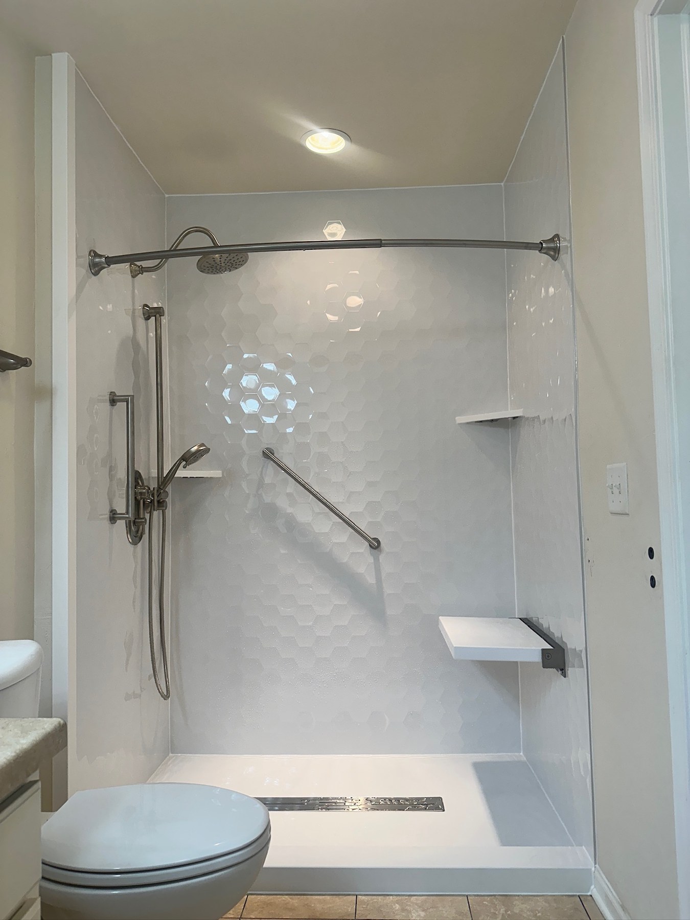 Walk in shower with Samuel Mueller Gloss White Hexagon walls, a brushed nickel rain shower head and handheld on a slide bar, grab bars, a white fold down seat, corner shelves and a linear drain installed by American Shower Company in Appleton, WI