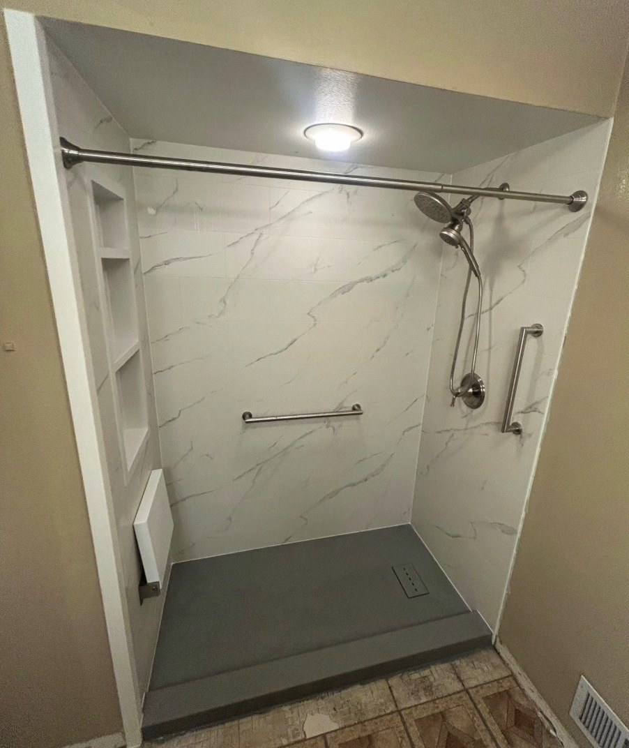 Walk in shower with white marble look walls, a recessed niche with shelves, a brushed nickel rain shower head and handheld, grab bars, a white fold down seat and a grey low threshold base installed by American Shower Company in a Wisconsin home