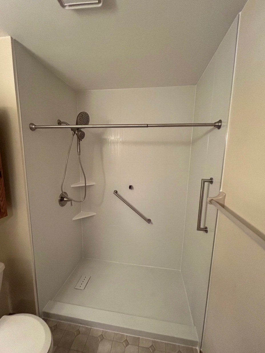 Walk in shower with smooth white walls, a brushed nickel rain shower head and handheld, corner shelves, grab bars and a white low threshold base installed by American Shower Company in a Wisconsin home