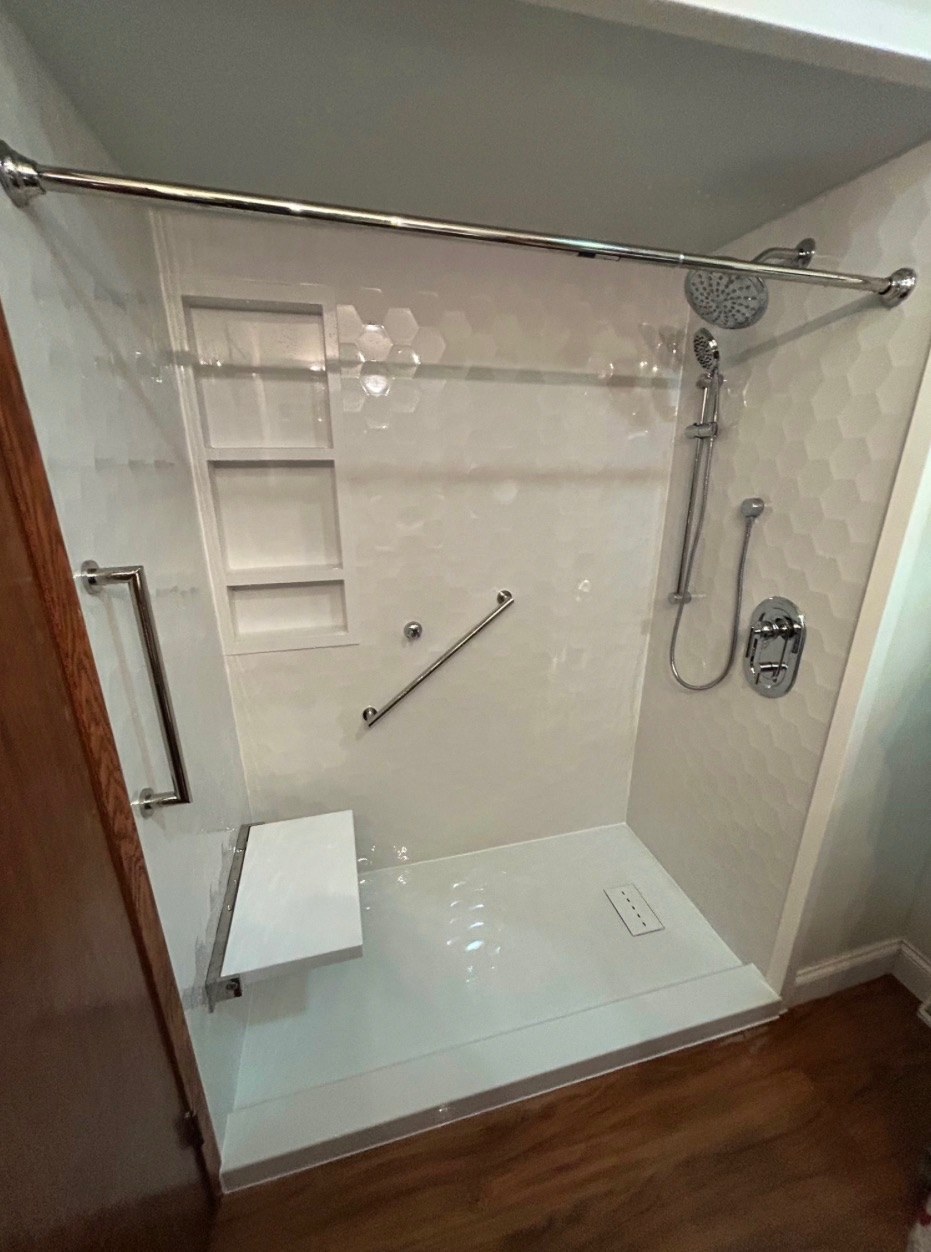 Walk in shower with Samuel Mueller Gloss White Hexagon walls, a three shelf recessed niche, a brushed nickel rain shower head and handheld on a slide bar, grab bars, a white fold down seat and a low threshold base installed by American Shower Company in a Wisconsin home